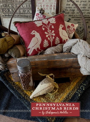 Decorative scene with a wooden basket containing pillows and yarn, featuring a cross stitch design of birds called Pennsylvania Christmas Birds by Shakespeare's Peddler