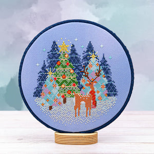 Cross-stitched design of reindeer and Christmas trees in a hoop on a medium blue aida fabric.